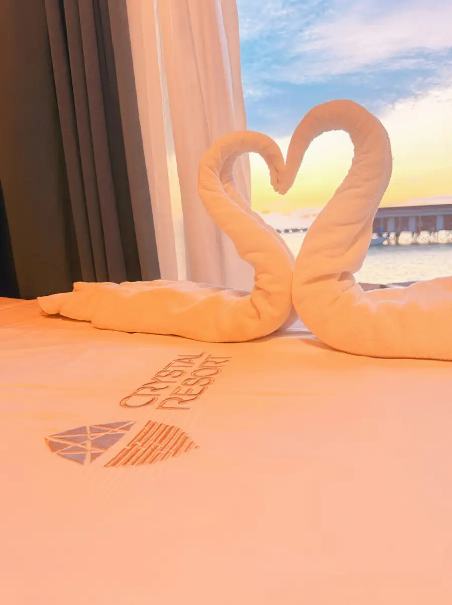 A cozy bed with towel art and resort logo next to glass doors revealing a vibrant sunset view over the Semporna islands at Crystal Resort